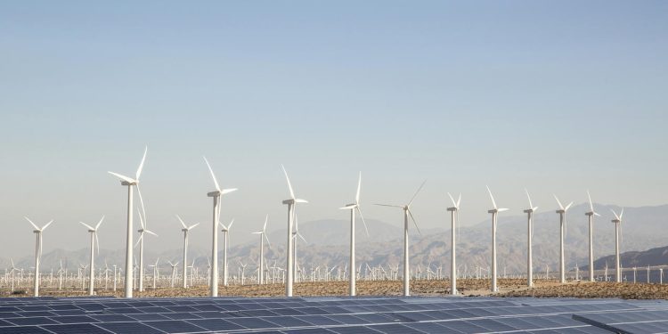 Many wind turbines and a large solar panel array in a desert valley, mountains in the distance and blue sky above. Palm Springs, California, USA